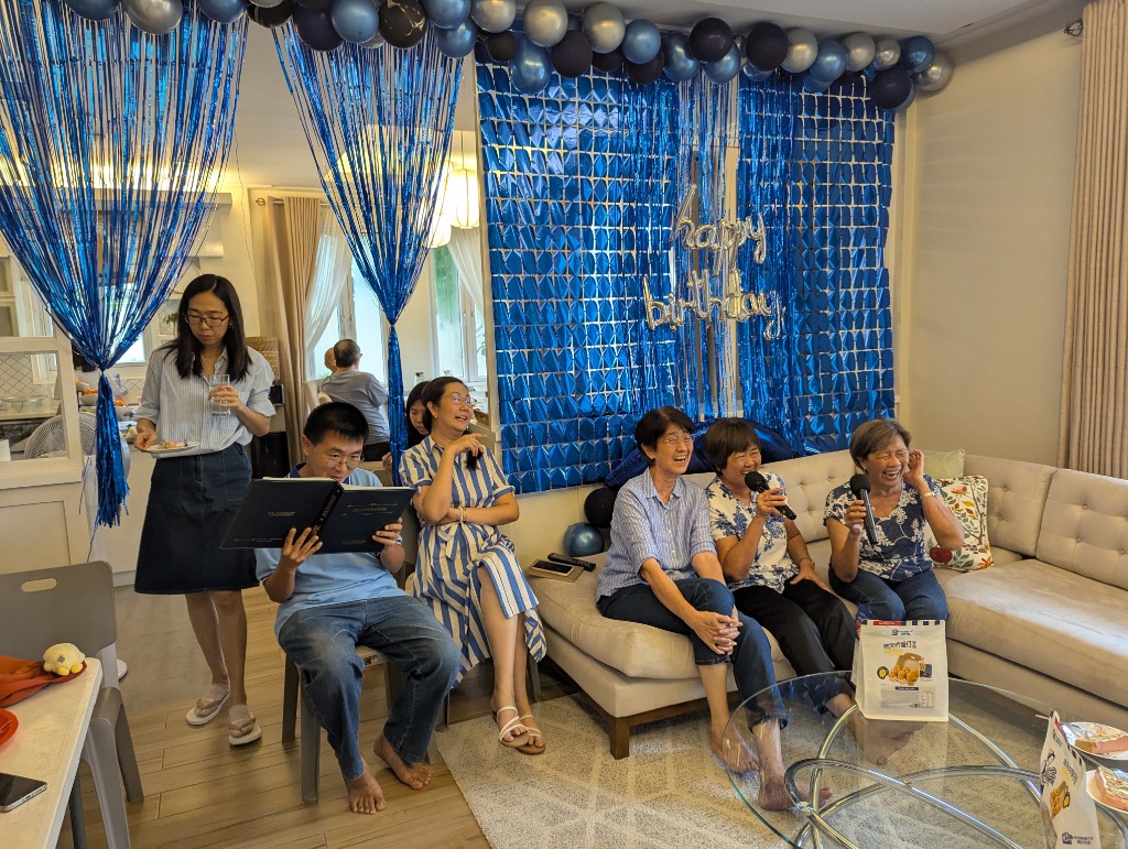 Wide shot of the full karaoke party living room with family laughing, singing, and the festive blue balloon backdrop