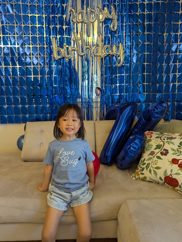 Little girl in Love Bug shirt posing in front of the blue happy birthday backdrop with 76 balloons